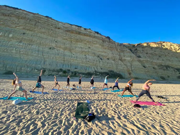 A group of people practicing Hatha yoga on yoga mats on a sandy beach in Lagos during a sunny morning.