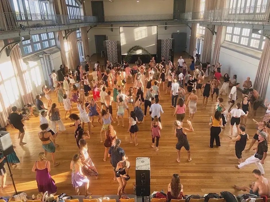 A wide-angle shot of a large, dimly lit hall in Lisbon filled with nearly 200 people dancing barefoot and moving freely during an Ecstatic Dance session.